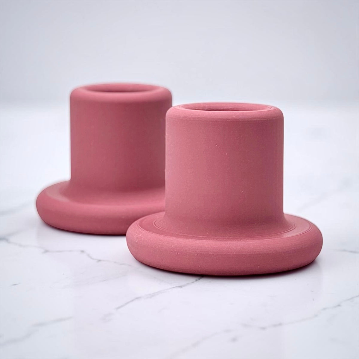 Two matte dusty pink silicone cup holders with a flared base, arranged on a white marble surface against a light gray background.