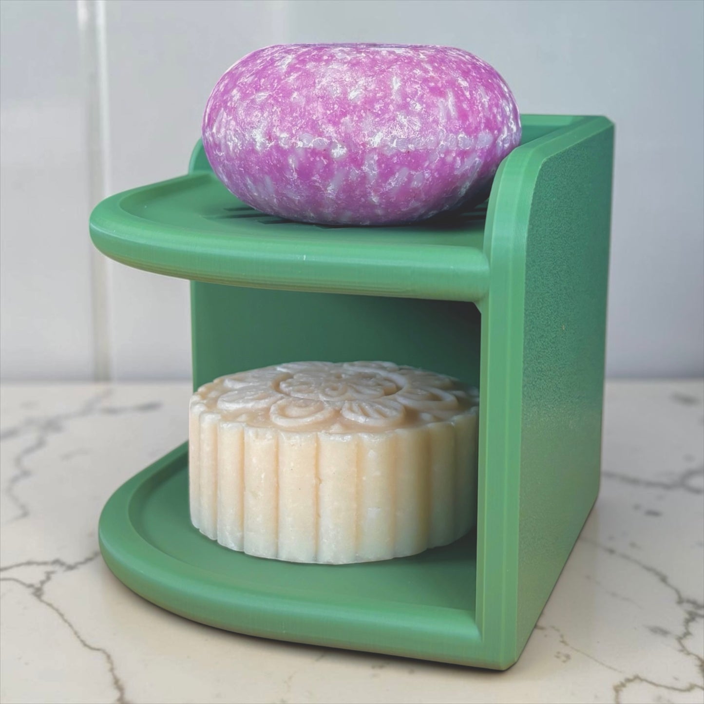 Olive Green soap holder with a purple and white speckled bar of soap on top and a white bar of soap below on a marble surface.