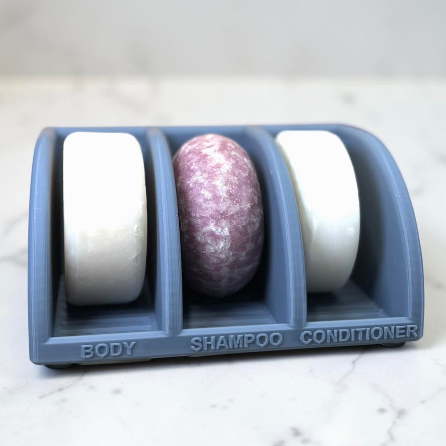 Three soap bars in a misty blue holder labeled 'Body', 'Shampoo', and 'Conditioner' on a marble surface.