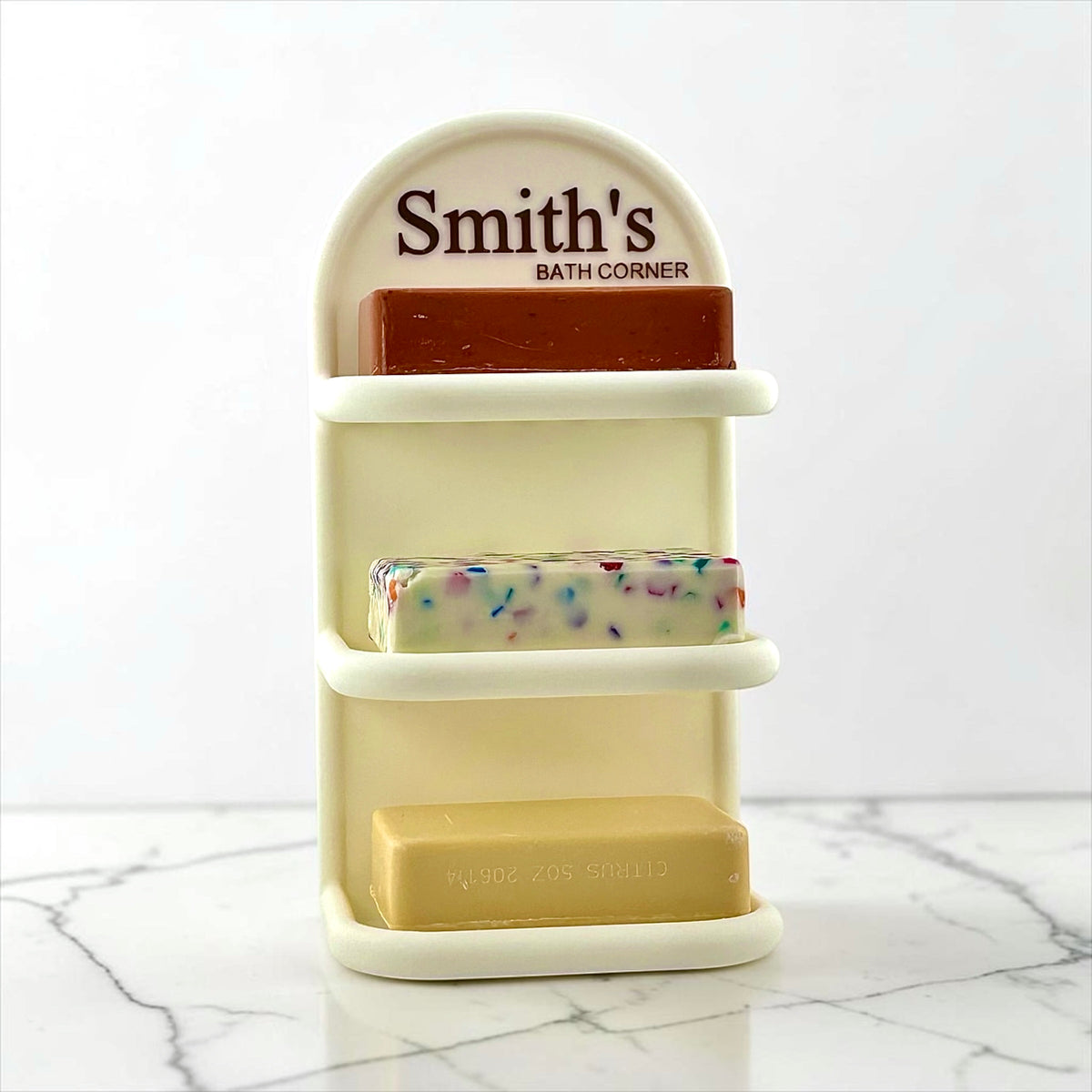A cream-colored three-tier soap holder with the personalized name 'Smith's BATH CORNER' printed on the top arch. The holder contains three bars of soap: a dark reddish-brown bar on top, a white bar with colorful flecks in the middle, and a beige bar on the bottom.
