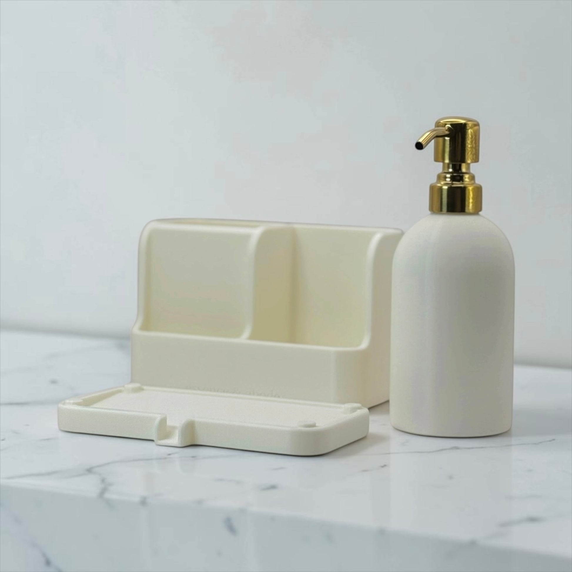 An off-white bathroom vanity set including a dual-compartment holder and a matching pump bottle with a gold pump dispenser, displayed on a white marble countertop against a light gray wall.