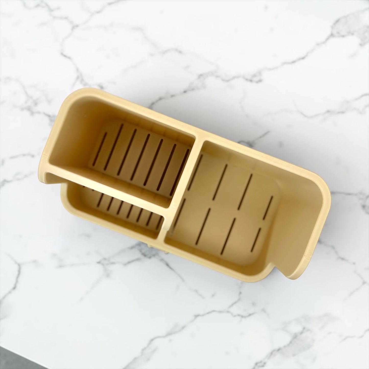 A beige plastic kitchen sink organizer tray with three compartments and drainage slots, shown from an overhead angle on a white marble surface.