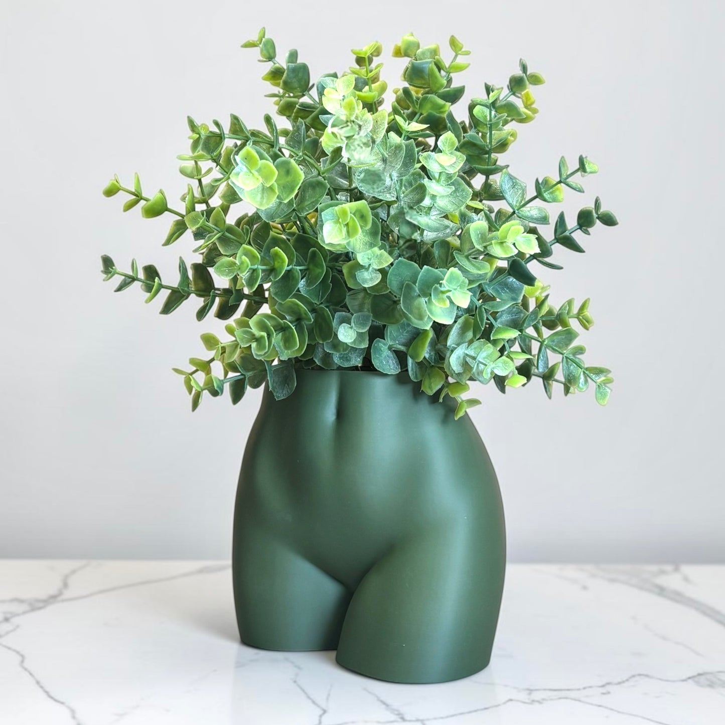 Dark Green vase shaped like a person's buttocks with a plant on a marble surface