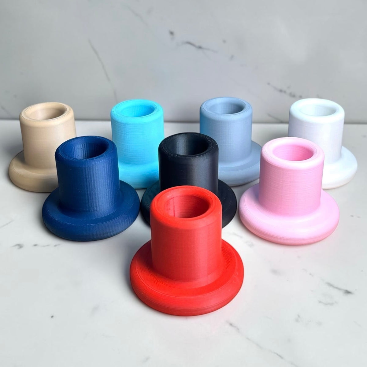 A set of eight 3D-printed cylindrical cup holders in various colors, including red, blue, black, light blue, beige, gray, pink, and white, arranged on a white marble surface.