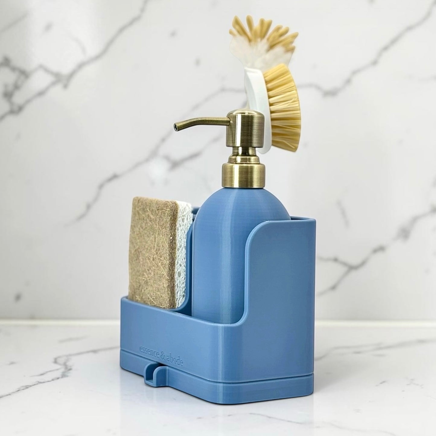A blue countertop soap dispenser organizer with a brass pump, a sponge, and a brush, set on a white marble surface against a white marble background.