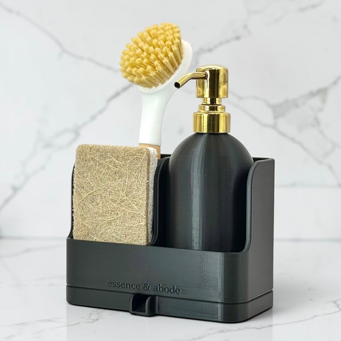 A black bathroom organizer tray holding a matte black pump bottle with a gold collar, a beige scrubber block, and a white-handled yellow bristle brush, set against a light marble background. The brand name 'essence & abode' is embossed on the front of the tray.