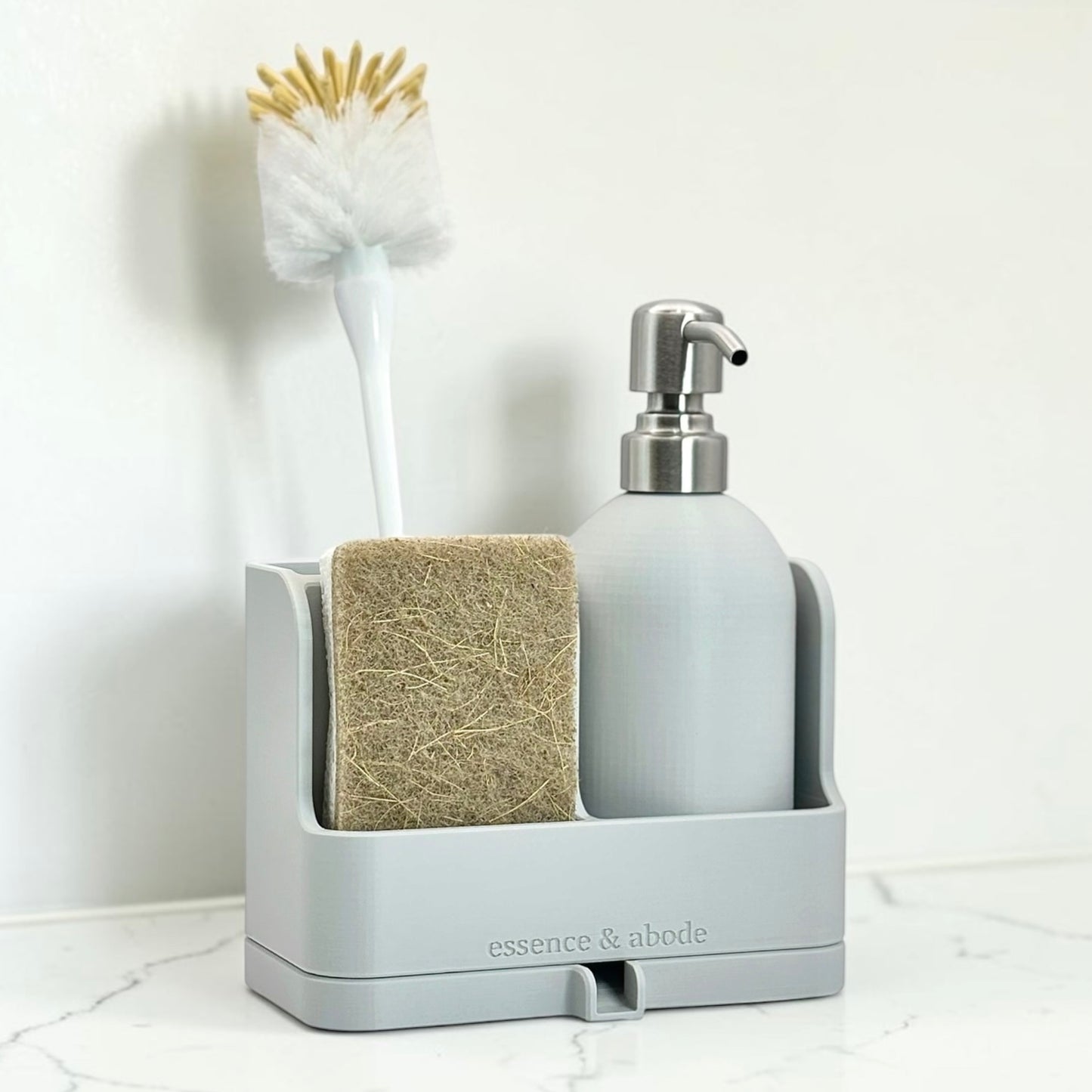 A light gray bathroom organizer tray holding a soap dispenser, sponge, and cleaning brush, on a white marble surface against a white wall. The brand name 'essence & abode' is printed on the front.