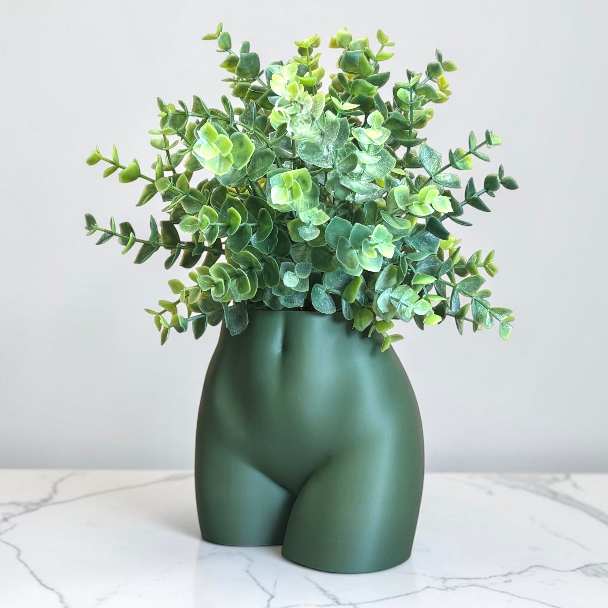 Green vase shaped like a person's buttocks with a plant on a marble surface