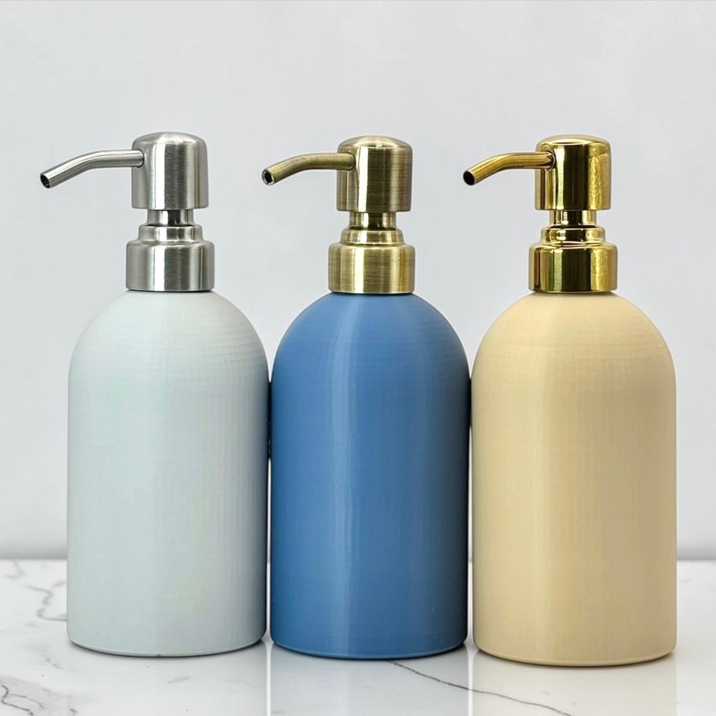 Three matte pump bottles in white, blue, and beige, each with a metallic pump dispenser, arranged side by side on a light marble surface against a white background.