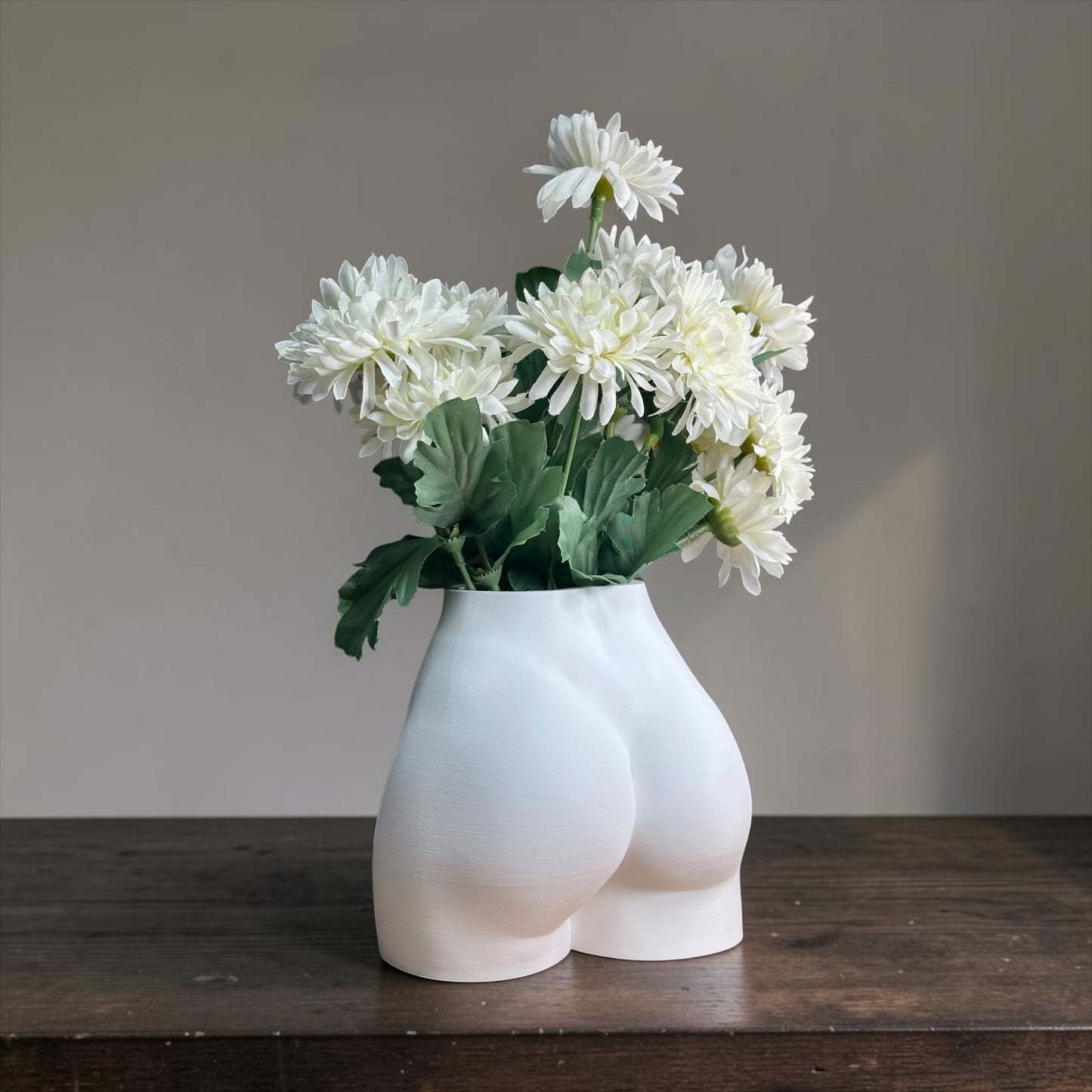 White vase shaped like a person's buttocks with white flowers on a wooden surface.