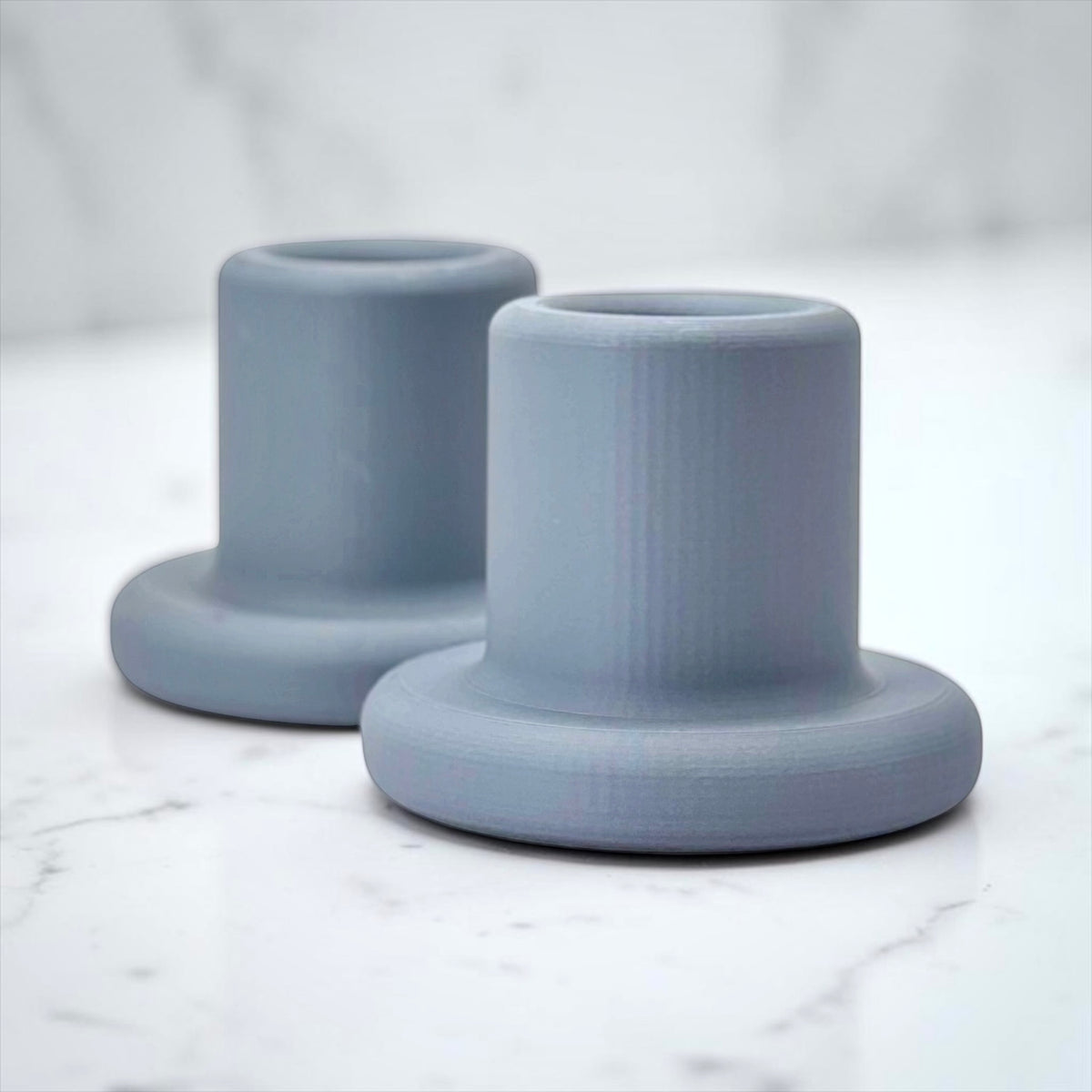 Two matte dusty blue cylindrical holders with flared bases, displayed on a white marble surface with a soft-focus marble background.