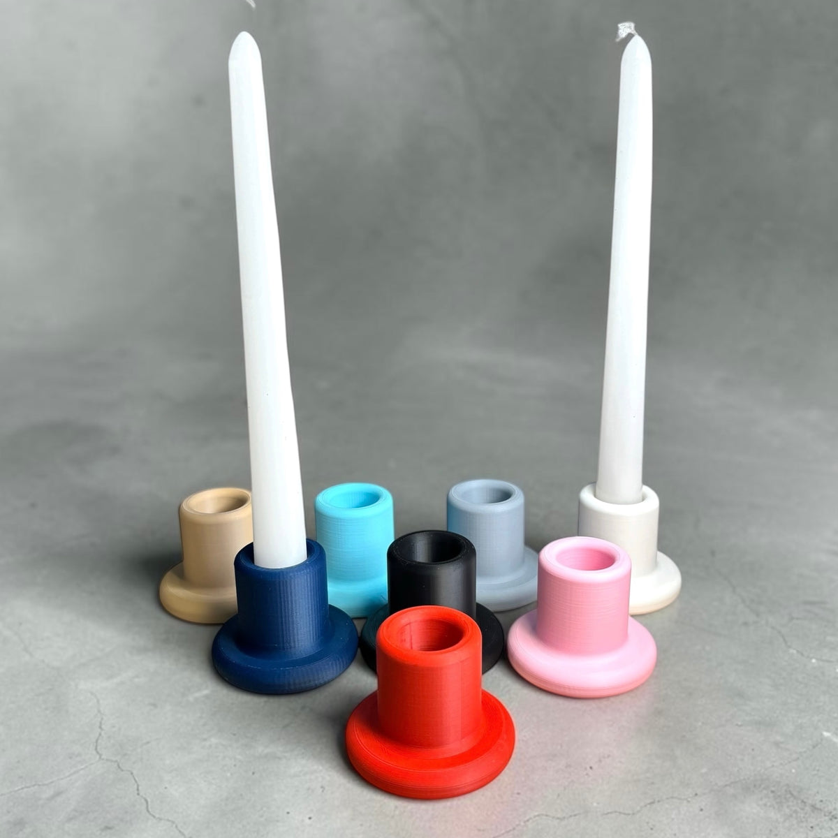 Two white taper candles standing in white holders, surrounded by multiple colored holder cups in beige, blue, black, gray, pink, and red on a gray textured background.