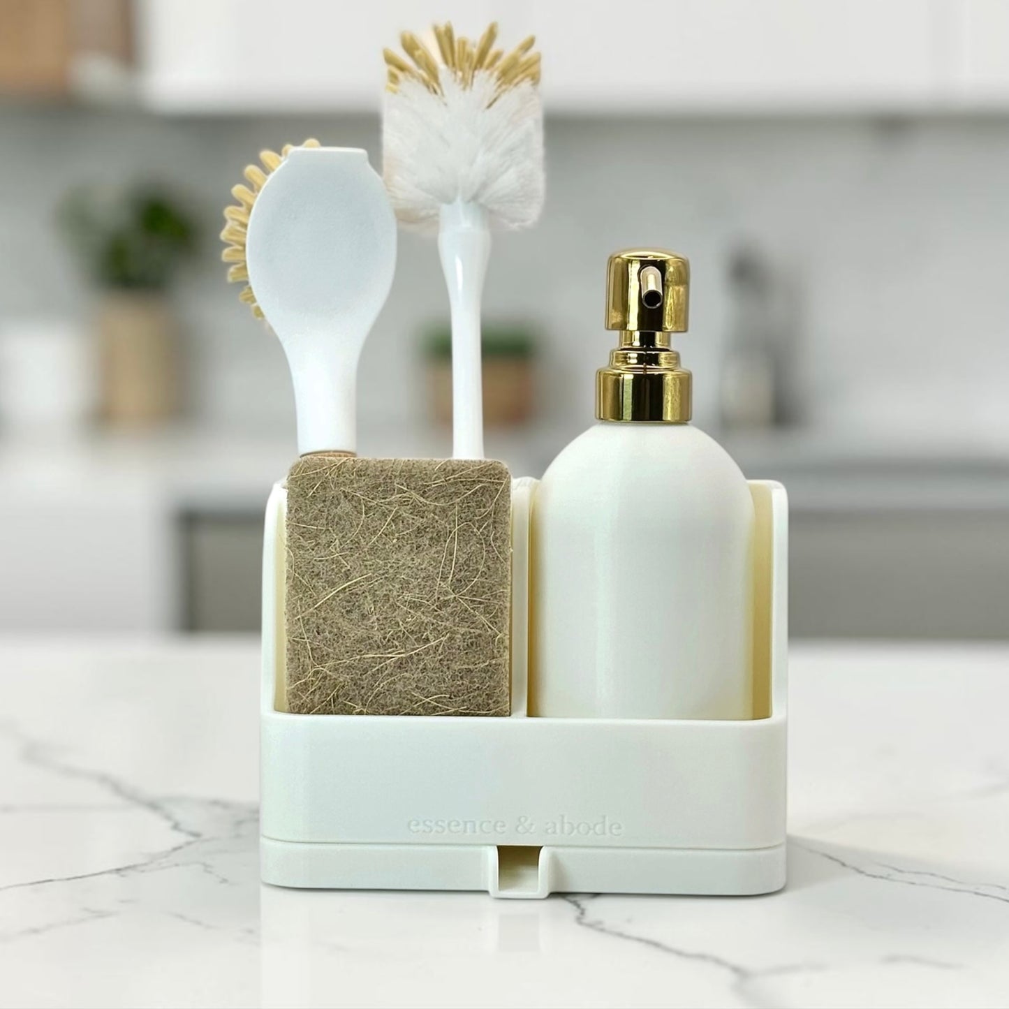 A white bathroom organizer set on a marble countertop, holding a white soap dispenser with a gold pump, a white sponge brush, and a white scrubber brush with natural bristles. A beige textured washcloth or sponge is tucked behind the dispenser.