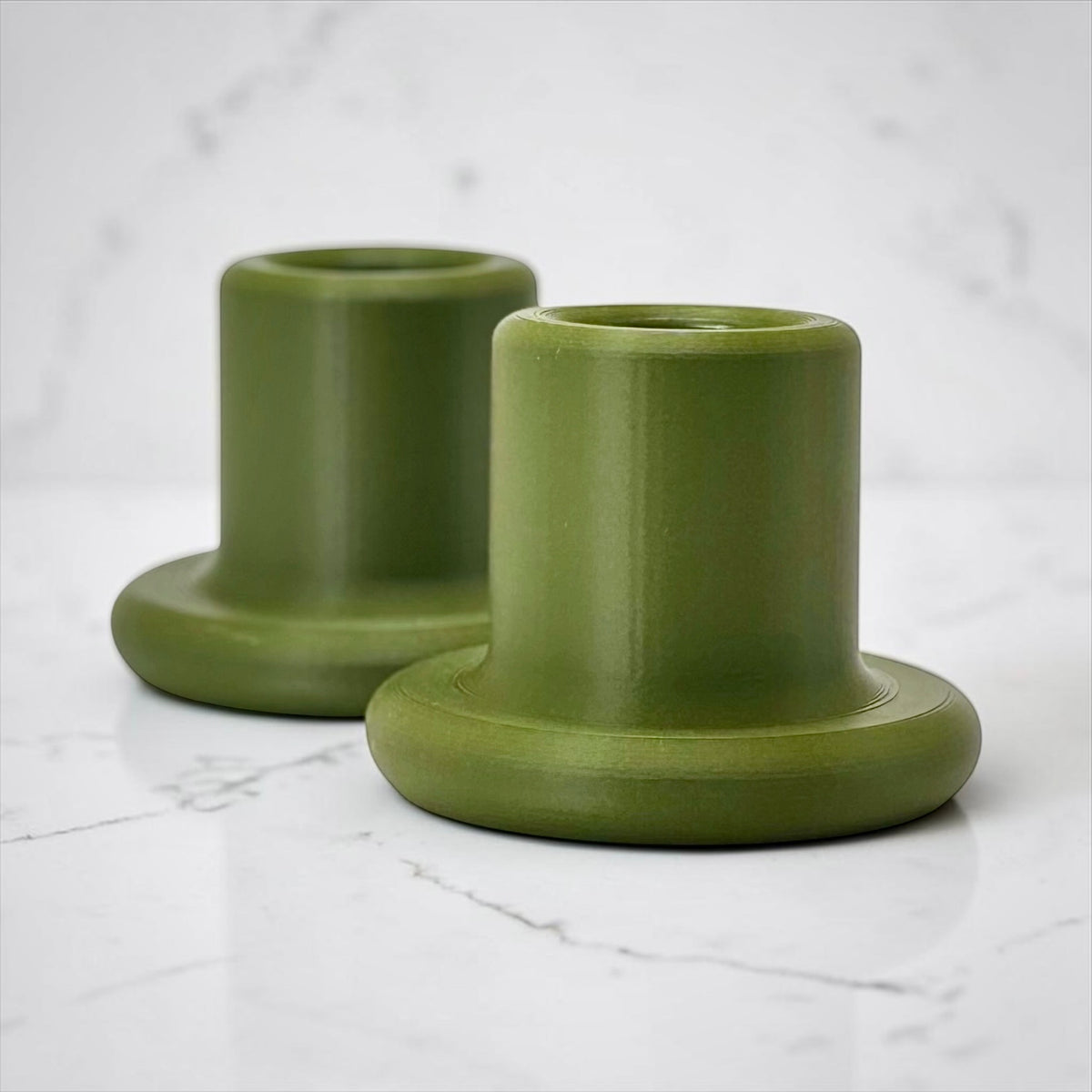 Two olive green ceramic candle holders with a matte finish, displayed on a white marble surface against a matching marble background.