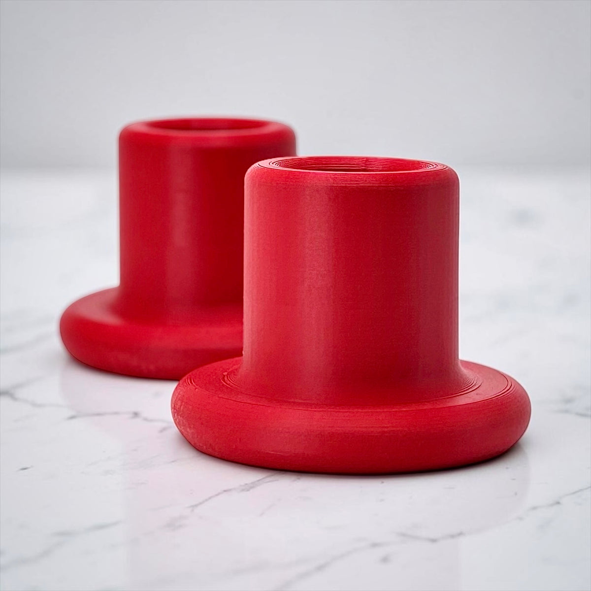 Two bright red cylindrical holder bases with flared circular bottoms, shown on a white marble surface with a soft gray background.