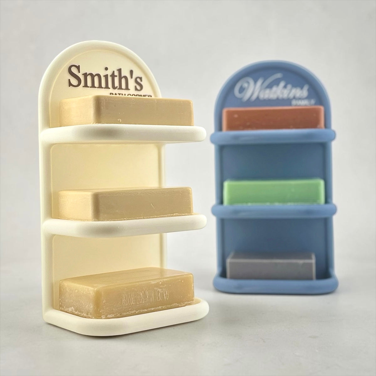 Two tower style soap dish organizers on a light gray background. The left organizer is off-white colored with the personalized name 'Smith's' printed on the top arch and holds three beige bars of soap.