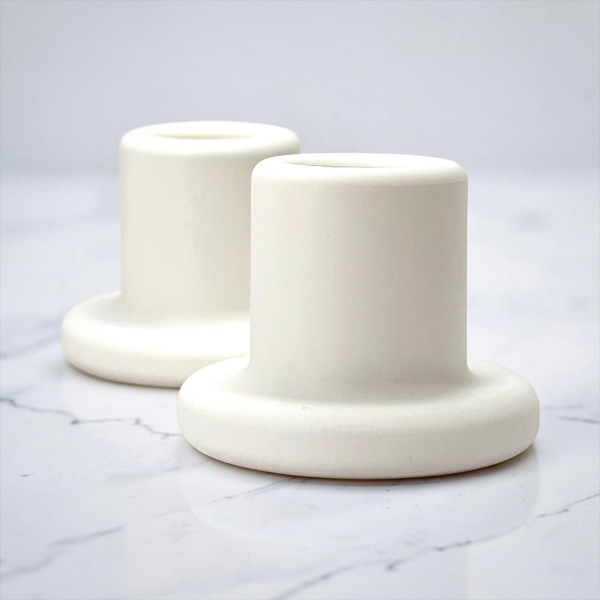 Two matte white cylindrical candle holders with flared bases, arranged on a light marble surface against a pale gray background.