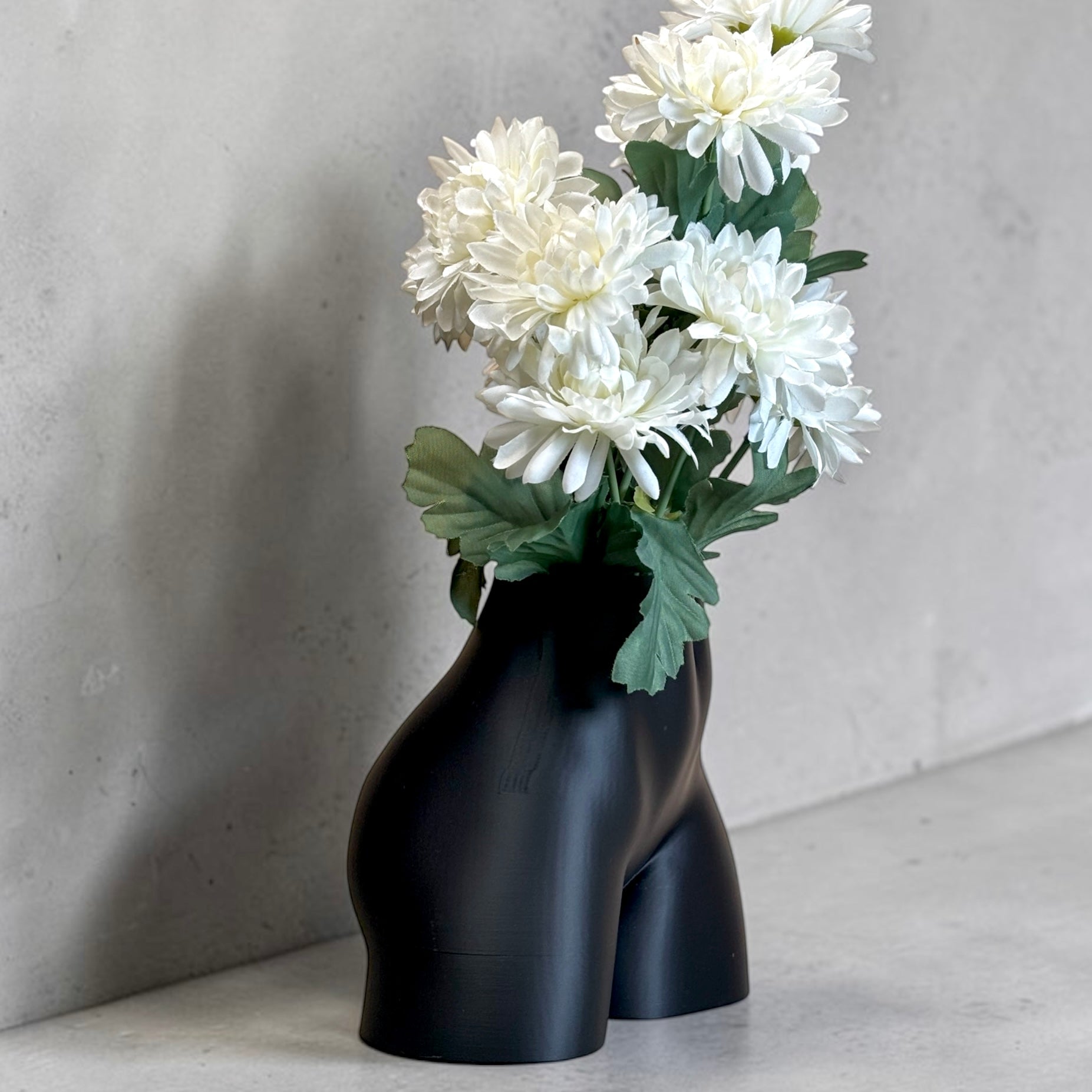 Black vase shaped like a pair of legs with white flowers against a gray background