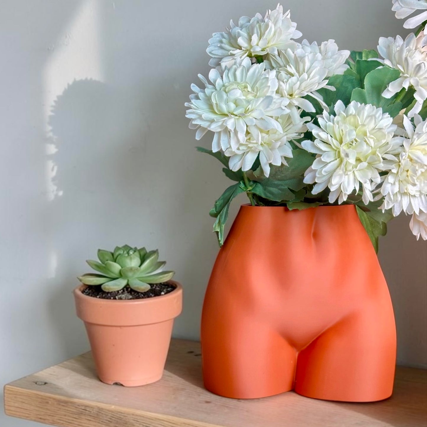 Terracotta Vase shaped like a woman's body with white flowers and a small potted plant on a wooden surface.