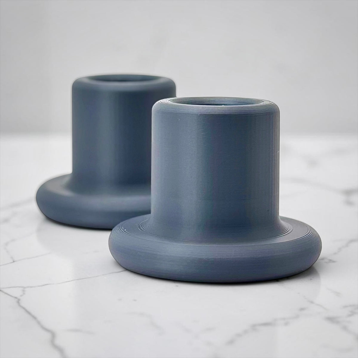 Two matte slate blue cylindrical candle holders with wide flared bases, arranged on a white marble surface against a light gray background.