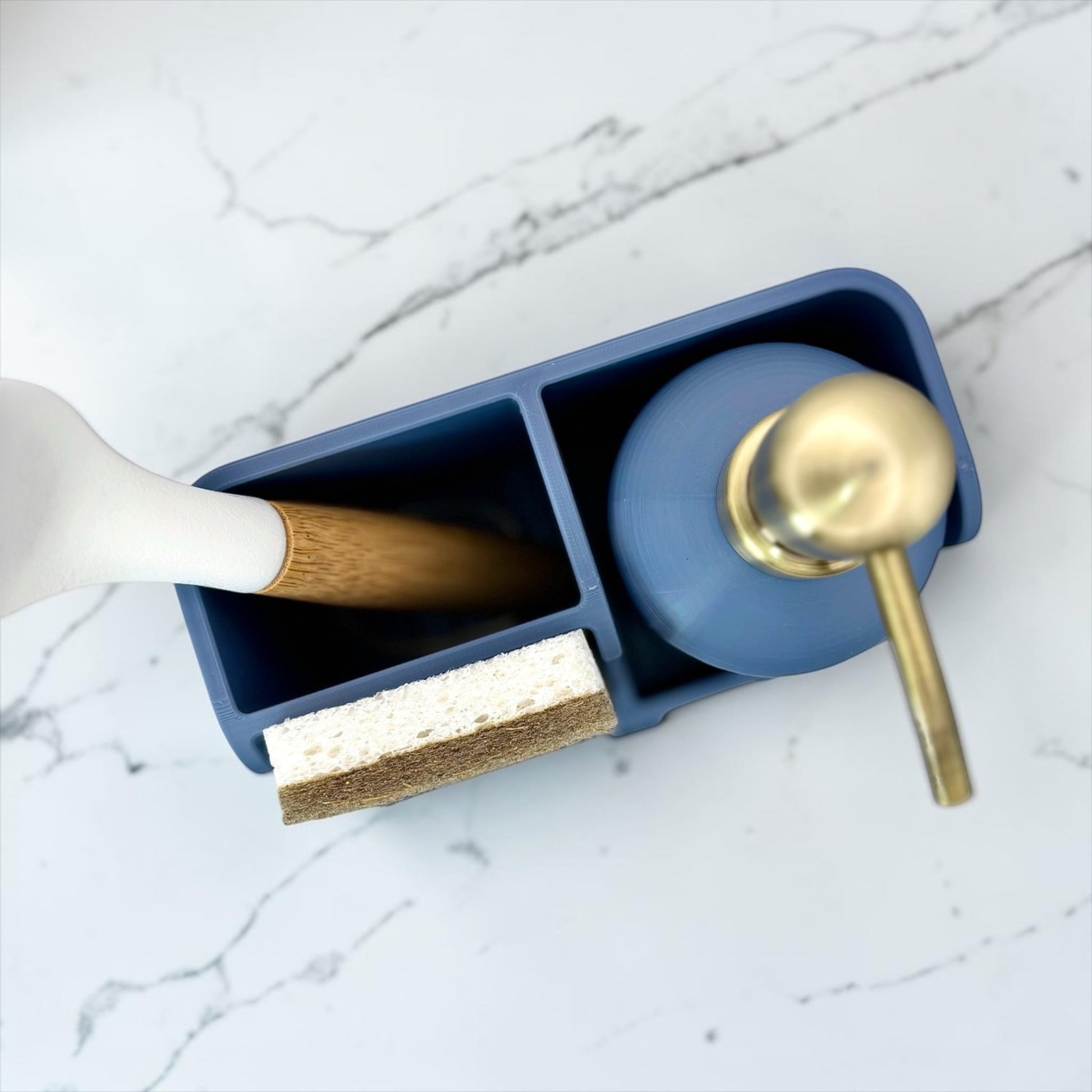 A blue bathroom organizer tray holding a wooden-handled toothbrush, a sponge, and a blue pump bottle with a gold-toned dispenser, set on a white marble surface.