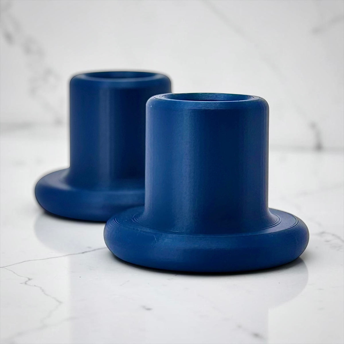 Two matte blue cylindrical holder bases with flared circular bottoms, photographed on a white marble-like surface with a soft white marble background.