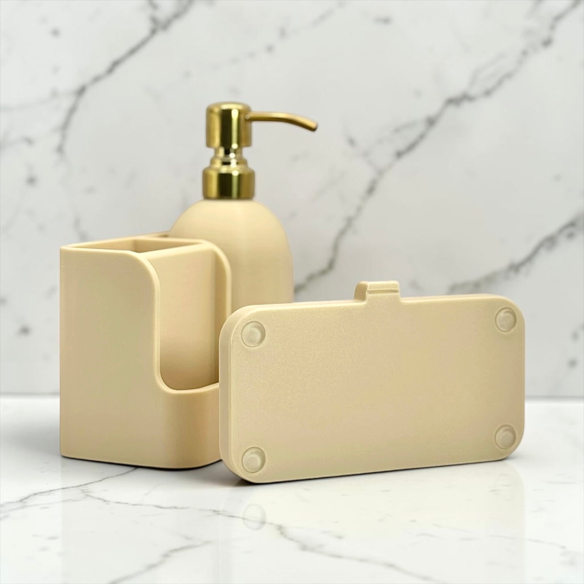 A beige bathroom accessory set on a white marble surface and backdrop, including a pump bottle with a gold-tone spout, a rectangular holder, and a small rectangular case.
