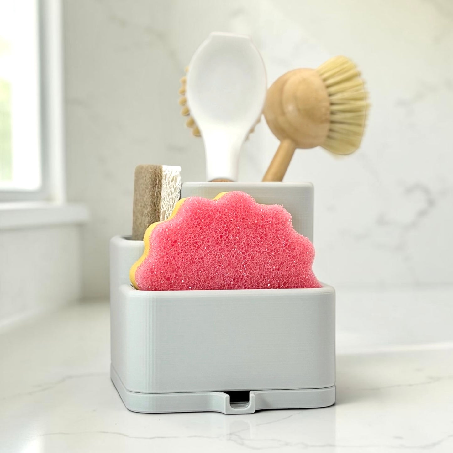 light gray caddy with a pink sponge, scrub brush, and wooden spoon on a white surface.