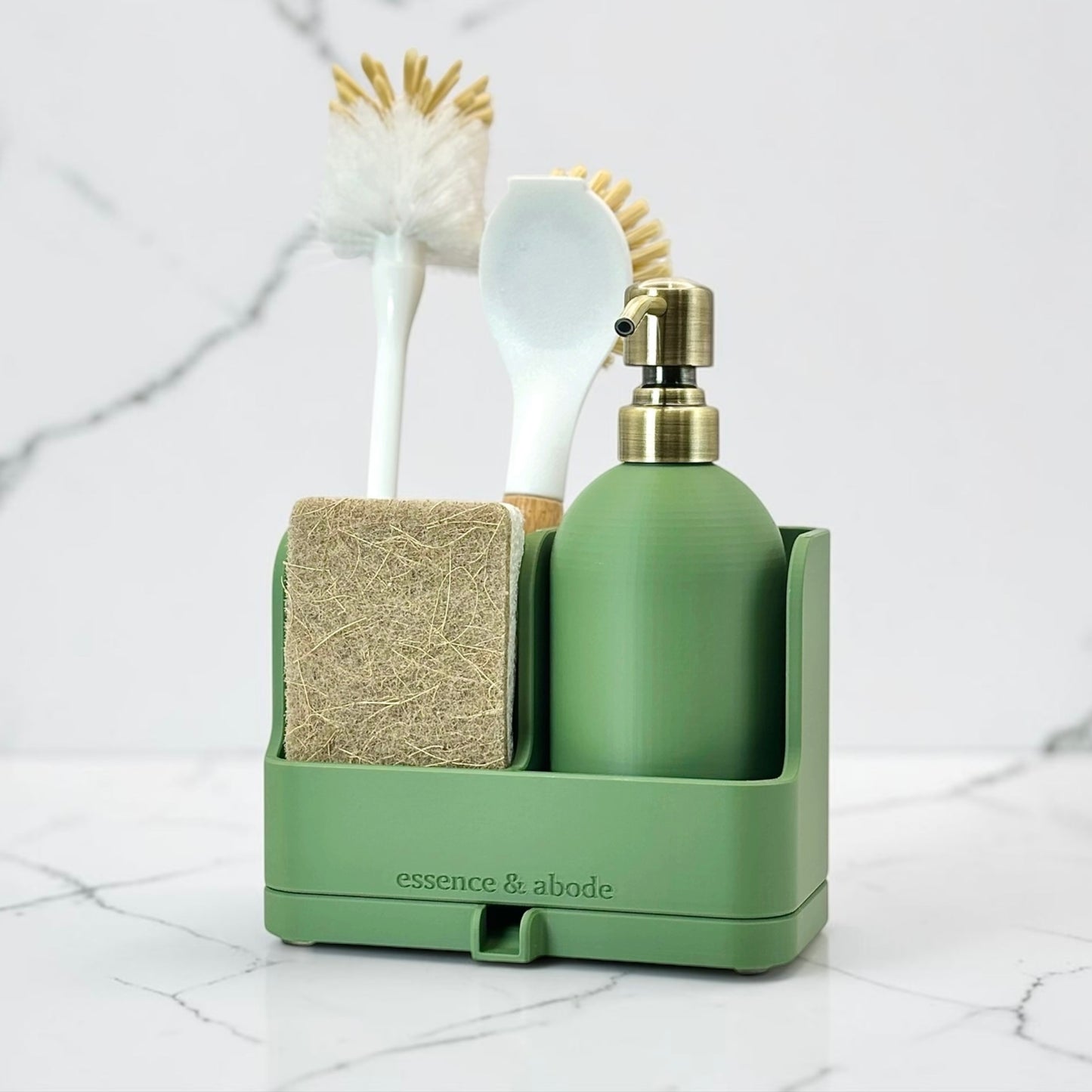 A sage green bathroom organizer set by essence & abode, containing a pump bottle, sponge, brush, and spatula, displayed on a white marble surface.