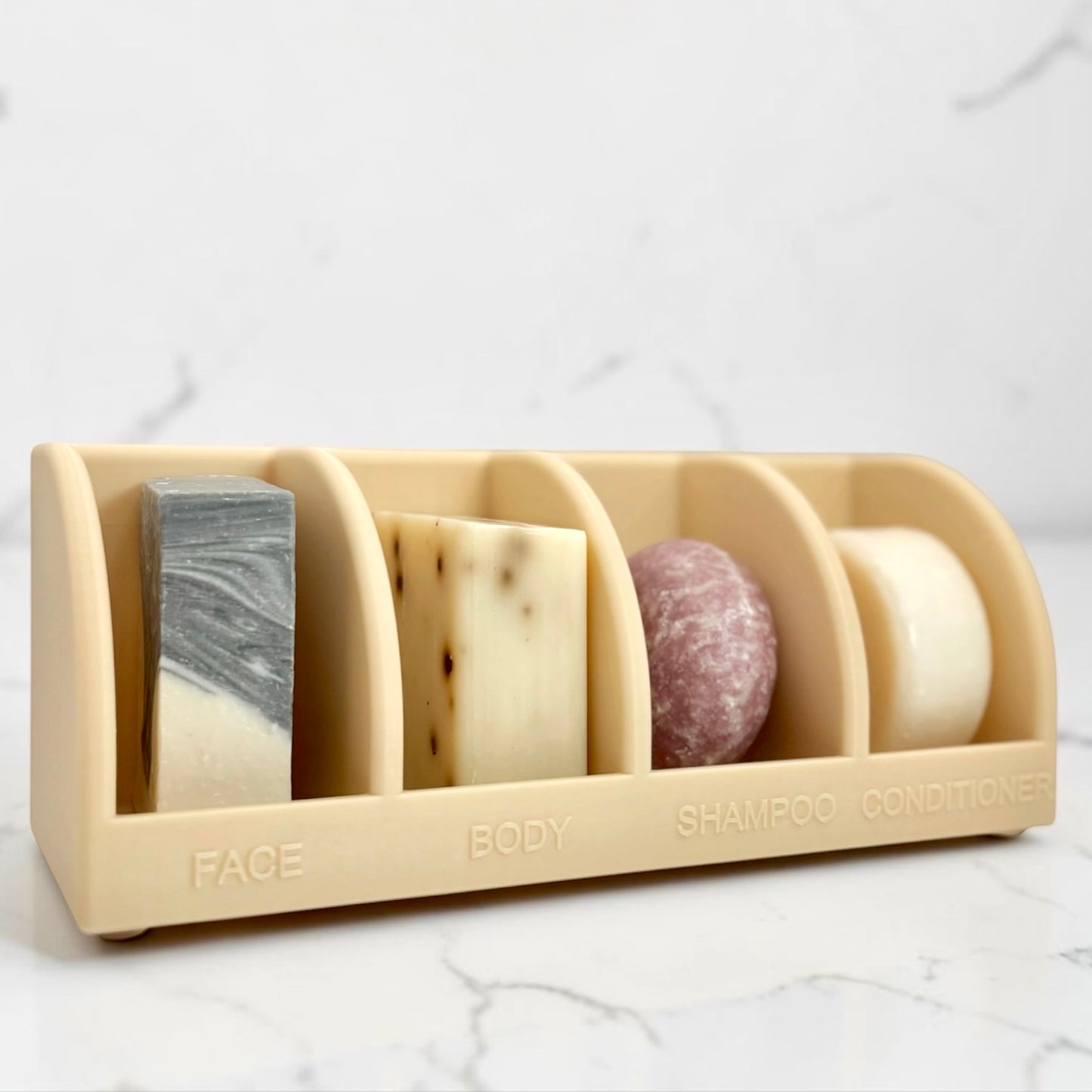 Beige soap holder with various soaps labeled 'Face', 'Body', 'Shampoo', and 'Conditioner' on a marble surface.