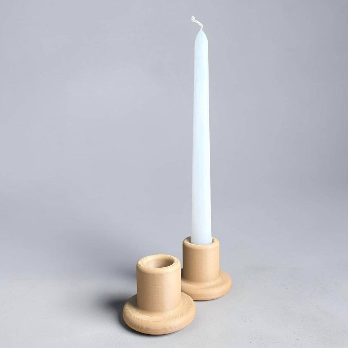 Two round beige ceramic candle holders on a light gray background, with a single white taper candle standing in the front holder.