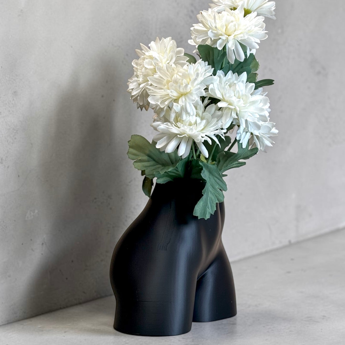 Black vase shaped like a women’s buttocks with white flowers against a gray background