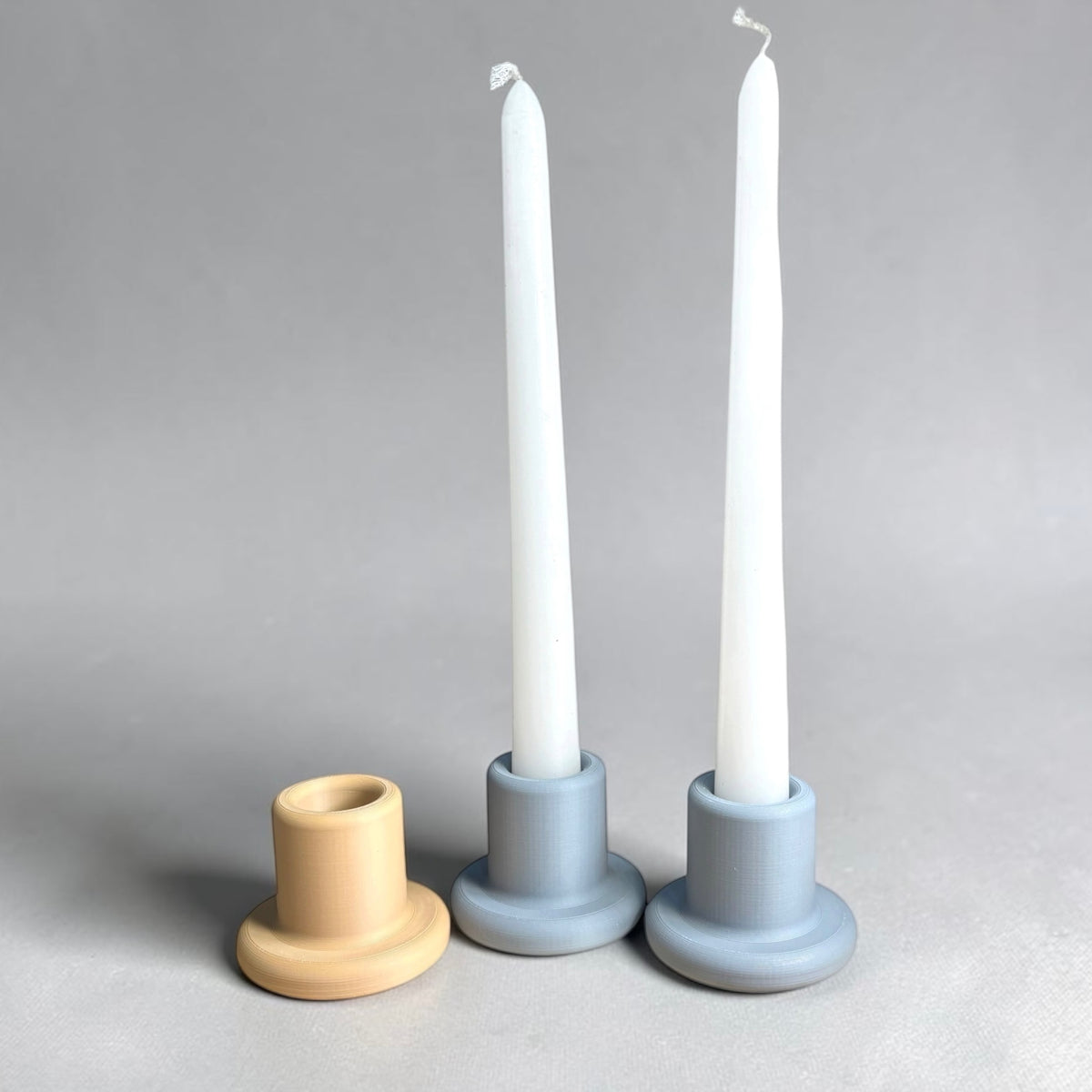 Three minimalist ceramic candle holders on a neutral gray background: one natural beige holder and two light blue-gray holders with white taper candles inserted.