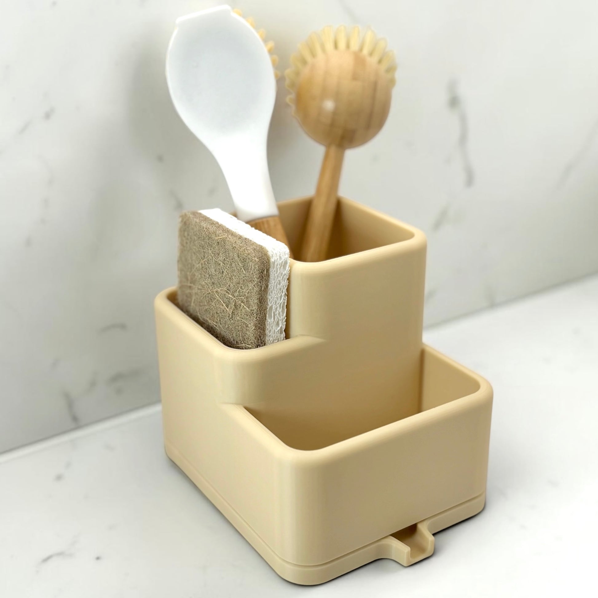 Beige sink caddy with a brush, sponge, and scrubber on a marble countertop.