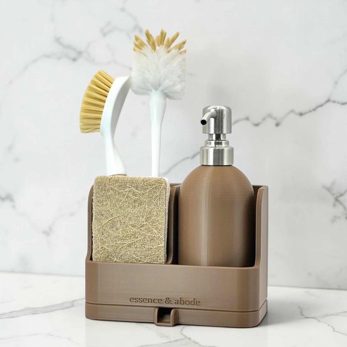 A brown bathroom organizer tray holding a soap dispenser, sponge, and two cleaning brushes, set on a white marble surface against a white marble wall. The tray is branded 'essence & abode'.