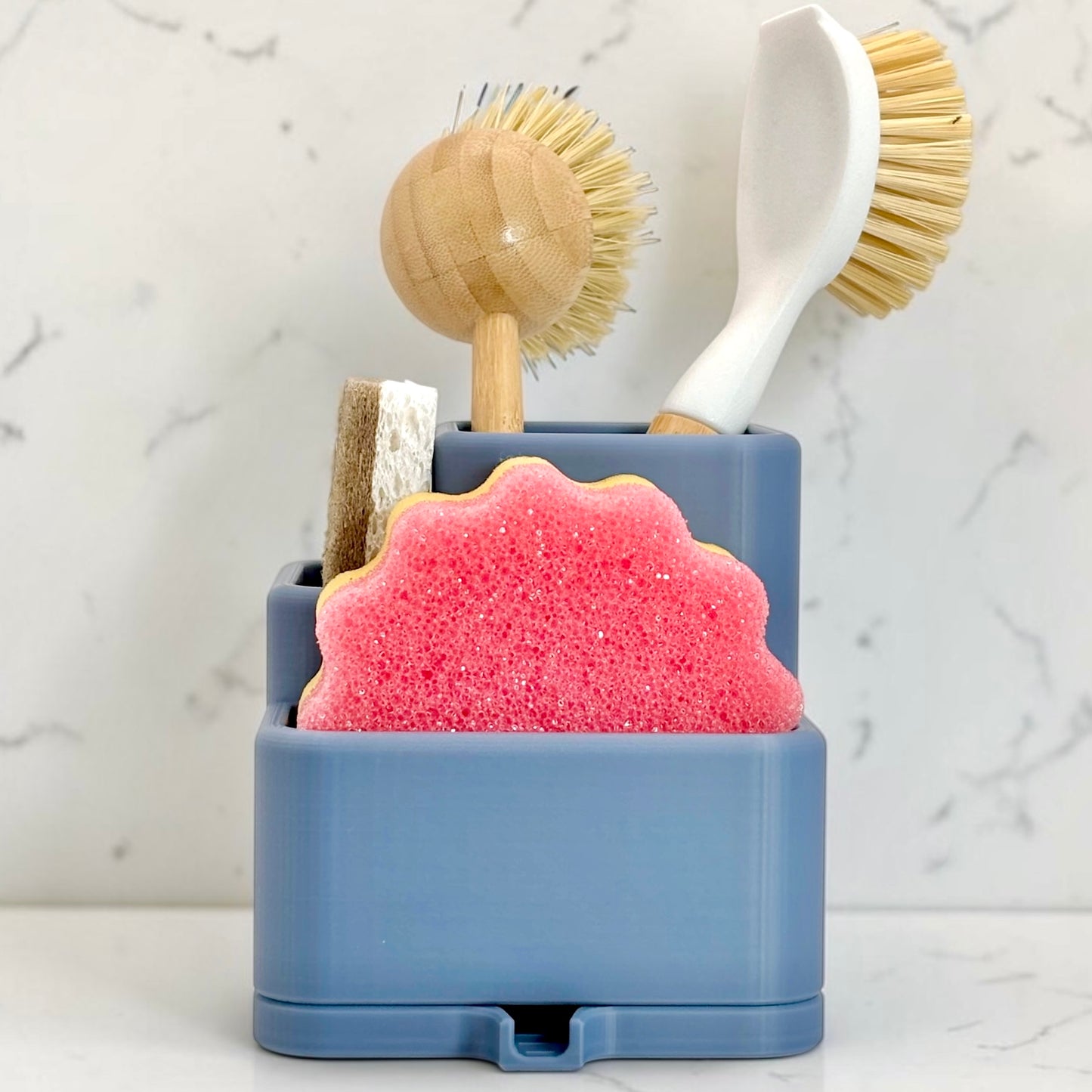 Blue holder with a pink sponge, wooden scrubber, and white brush against a marble background