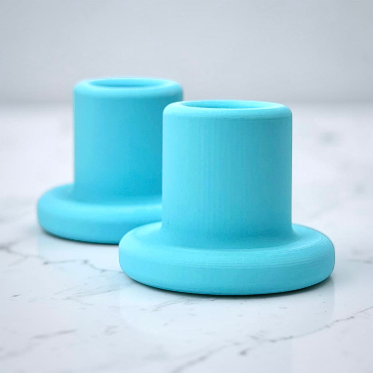 Two light blue 3D-printed cylindrical holders with flat circular bases, displayed on a white marble surface with a soft gray background.