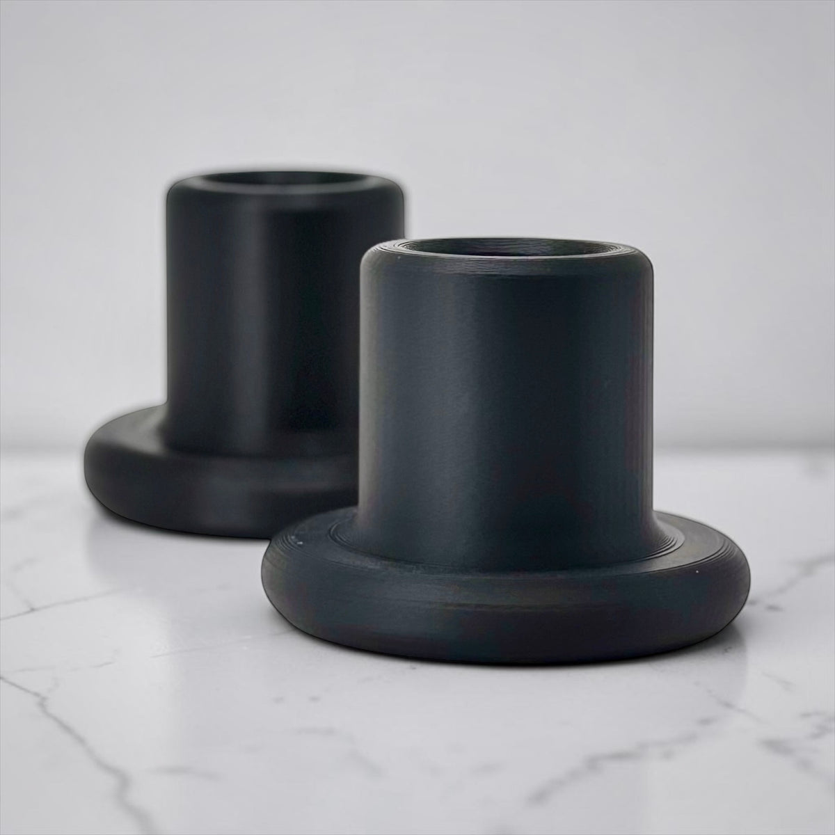 Two matte black cylindrical candle holders with flared bases, arranged on a white marble surface against a light gray background.
