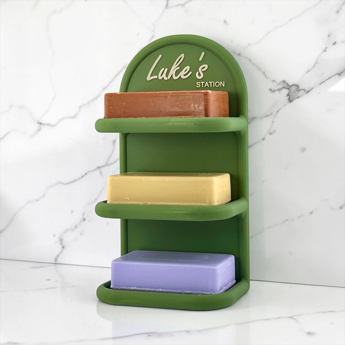 A green three-tier soap dish holder with an arched backplate printed with 'Luke's STATION' in white script, shown holding three rectangular bars of soap on a white marble surface against a white marble wall.