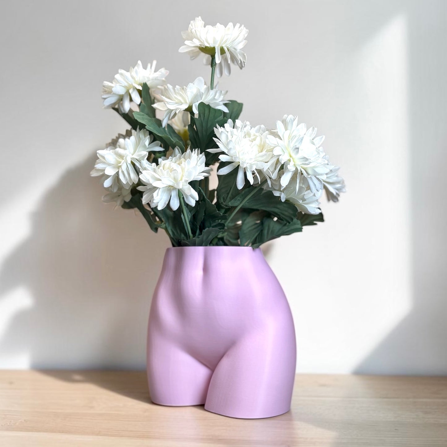 Light pink vase shaped like a women’s curvy torso with white flowers on a wooden surface