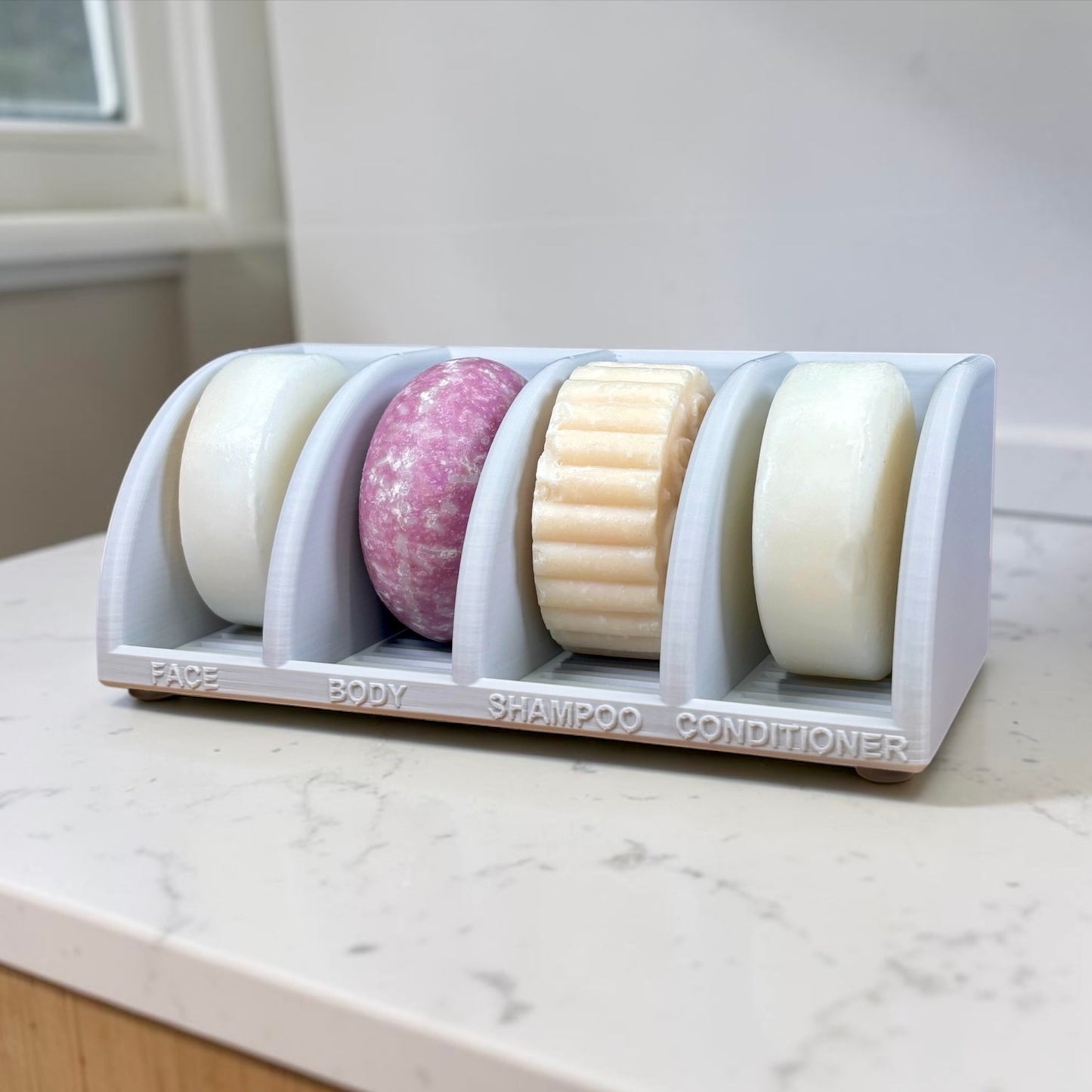 Soap holder with four soap bars labeled 'Face', 'Body', 'Shampoo', and 'Conditioner' on a marble countertop.
