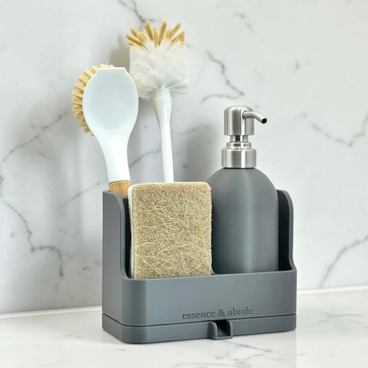 A gray bathroom organizer tray holding a soap dispenser, sponge, scrubber, and brush, set against a white marble background. The brand name 'essence & abode' is printed on the front of the tray.