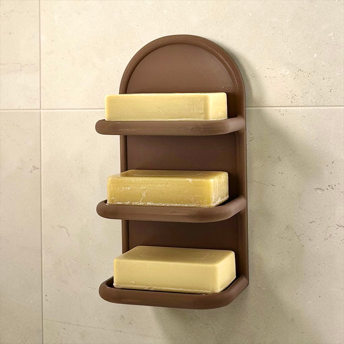 A three-tier brown plastic wall-mounted soap holder with bars of yellow soap, shown against a beige tiled wall.
