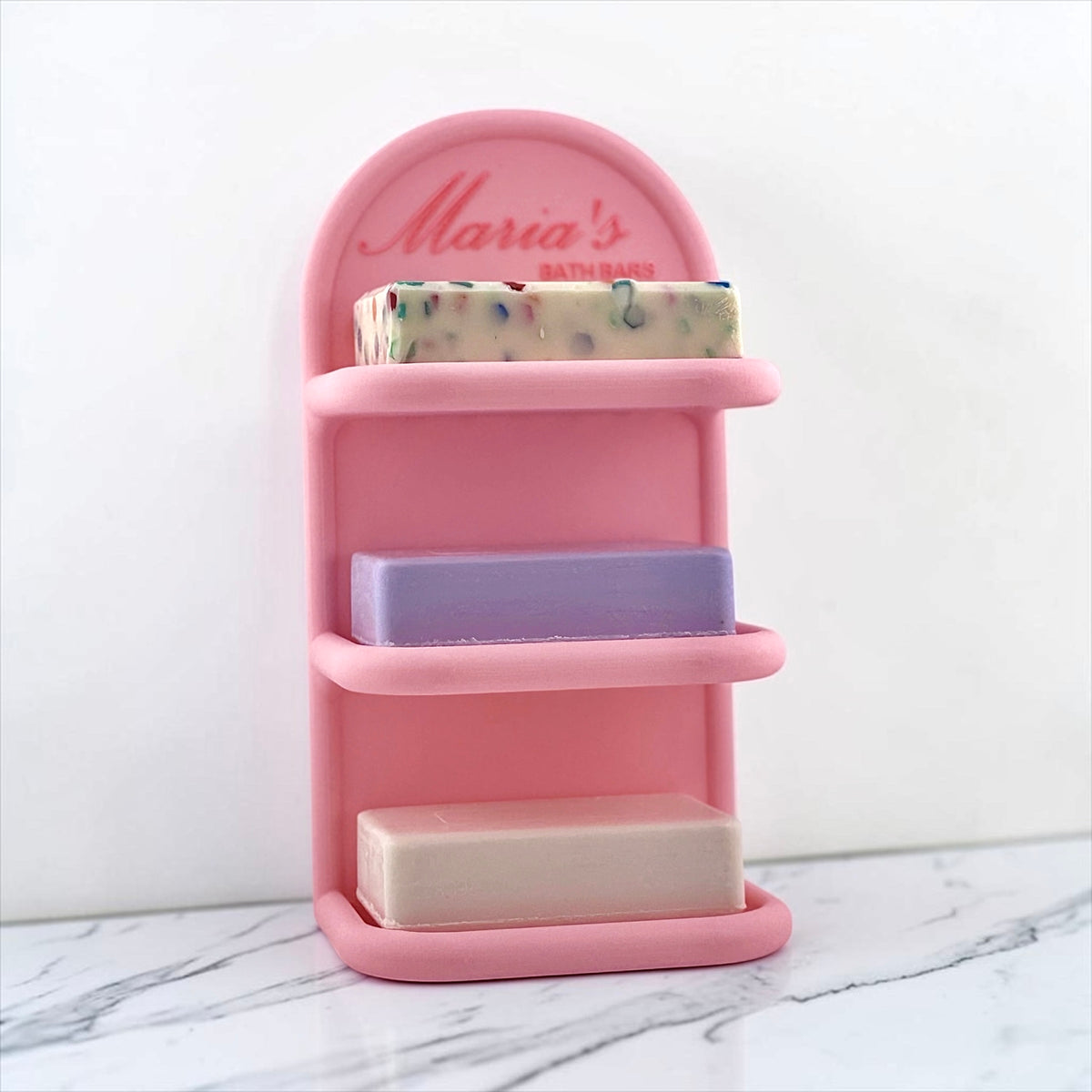 A pink three-tier soap holder with a curved backplate reading 'Maria's BATHSOAPS' in red script, shown holding three bars of soap on a white marble surface against a white wall.