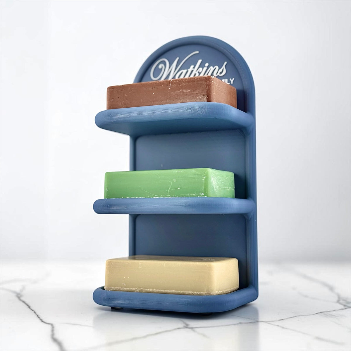 A blue three-tier soap holder with the personalized name 'Watkins Family' printed in white on the back panel, holding three rectangular bars of soap in brown, green, and cream, set against a white marble surface and background.