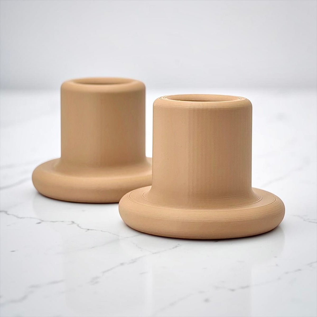 Two minimalist beige ceramic candle holders with a flared base and cylindrical top, displayed on a white marble surface against a light gray background.
