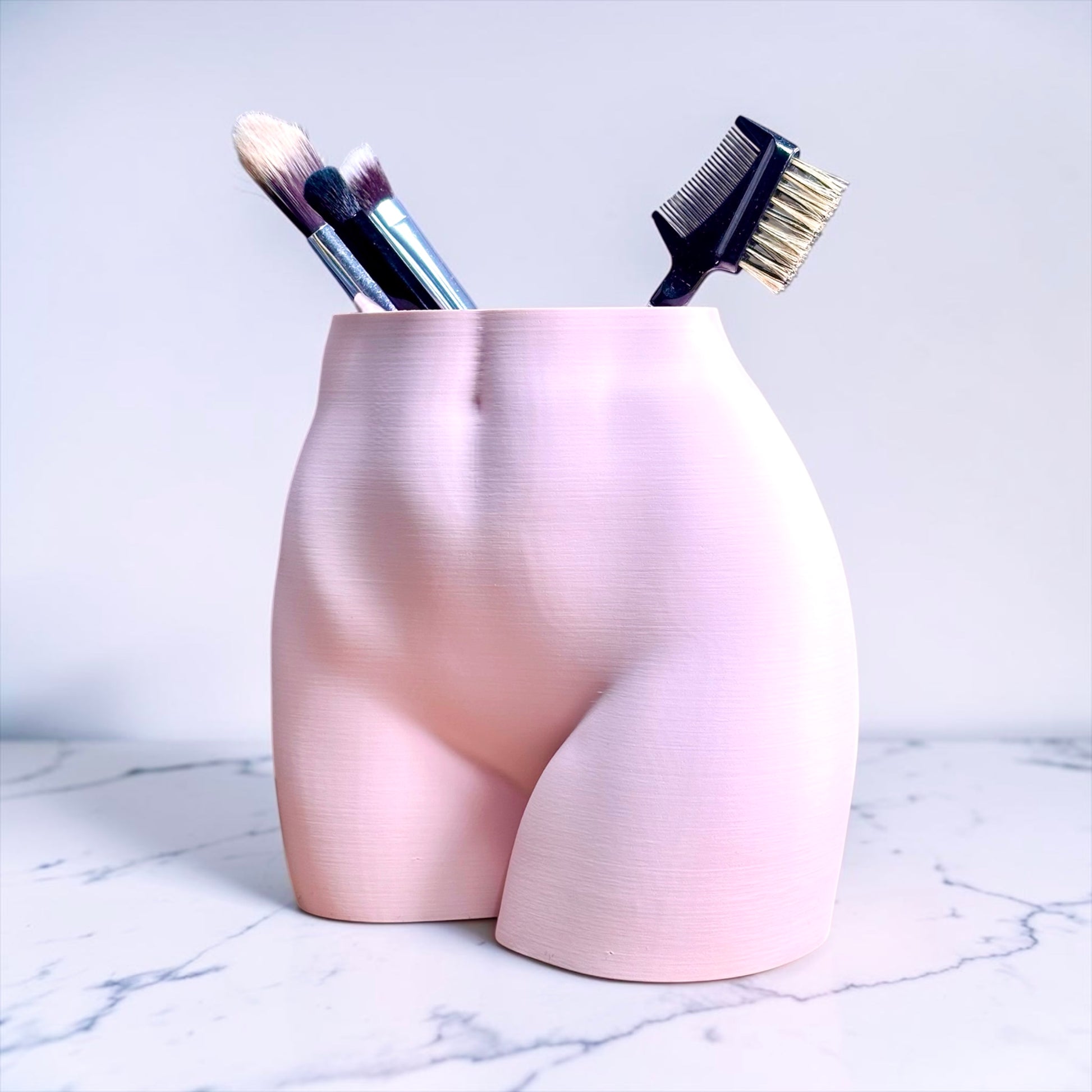 Pink plant-based plastic sexy butt shaped holder with makeup brushes on a marble surface