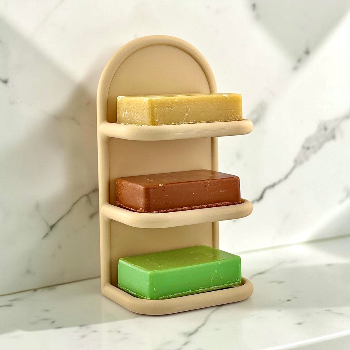 A three-tier beige soap holder filled with three rectangular bars of soap in yellow, brown, and green, set against a white marble background.