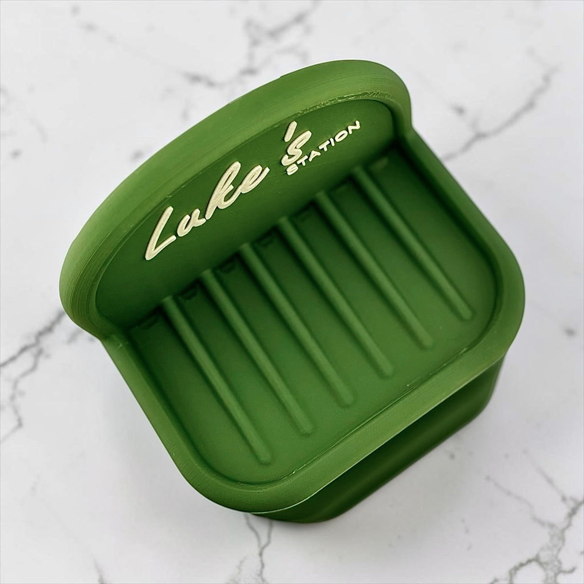An olive green soap holder, featuring the personalized 'Luke Station' printed in white on the front. set on a white marble surface.