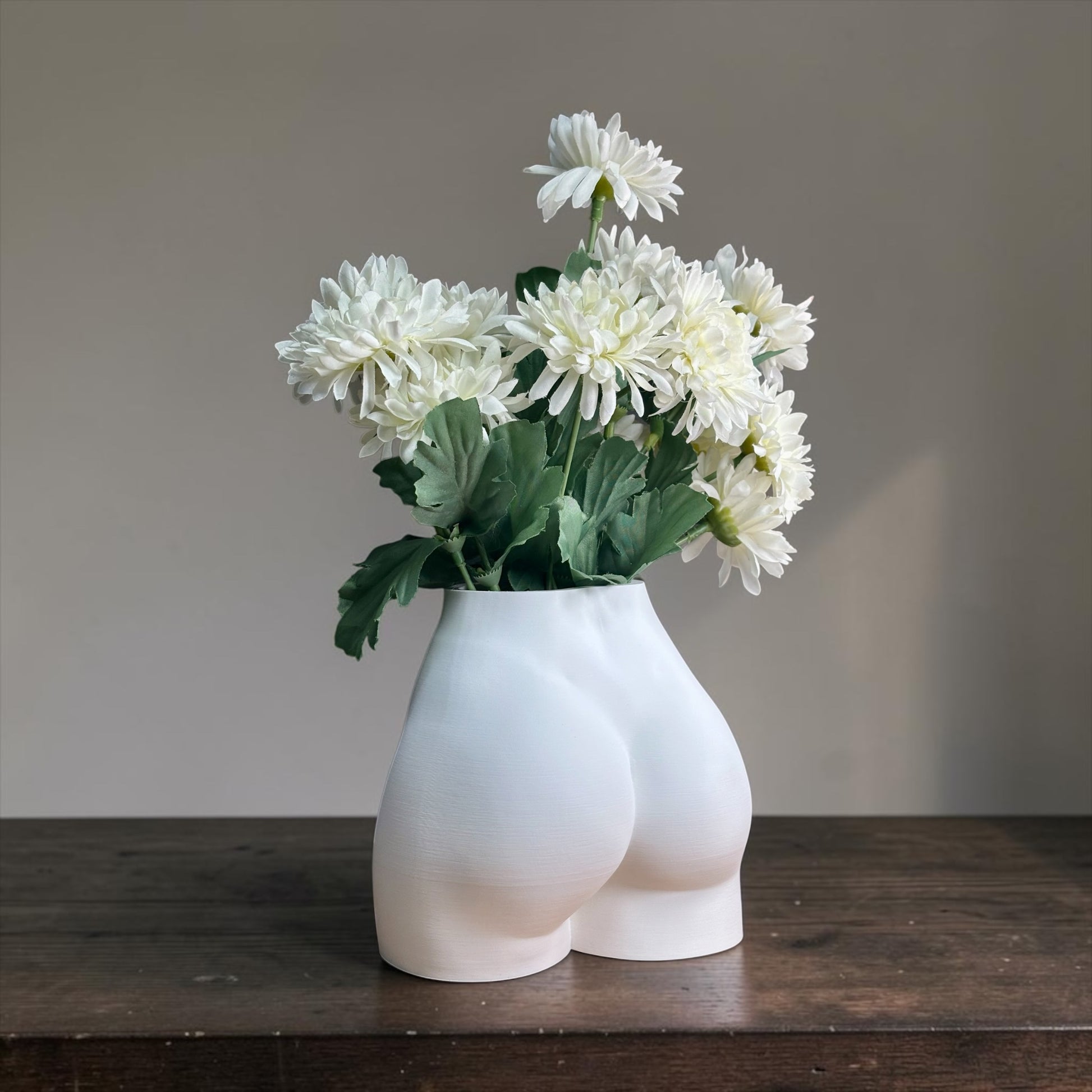White vase shaped like a person's buttocks with white flowers on a wooden surface.